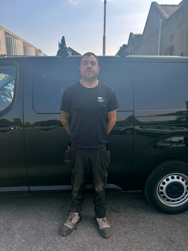 Image depicts Matt Brooks, Senior Supervisor at Woodward Group, standing in front of a Woodward Group black van. Matt's T Shirt has the Woodward Group logo on it.